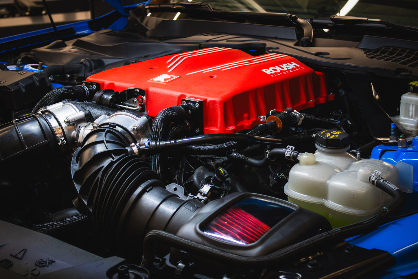 A powerful car engine featuring a striking red Roush Mustang Supercharger, with its 810 horsepower potential on display, showcases visible motor components intertwined with an array of pipes and wires.