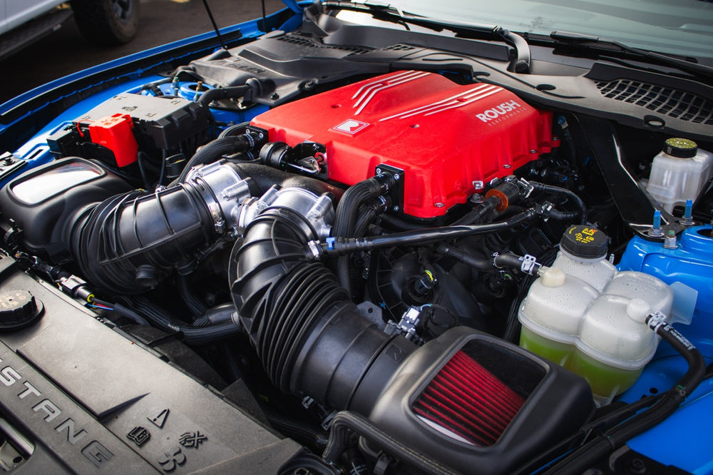 Close-up of a car engine with a red cover featuring the Roush logo. The supercharger kit powers this beast to an impressive 740 horsepower. Black hoses and various engine components are visible, with the bottom of the hood showcasing 