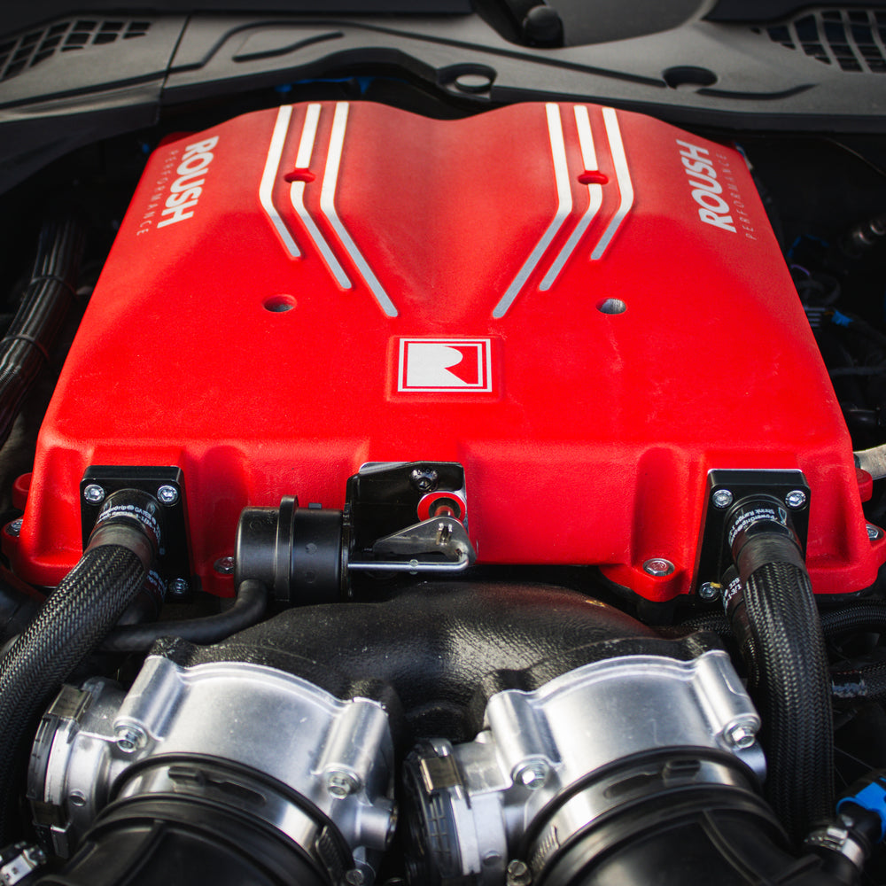 
                  
                    Close-up of a red Roush S650 Mustang engine cover with visible hoses and components under a car hood, showcasing its impressive 740 horsepower Supercharger Kit.
                  
                