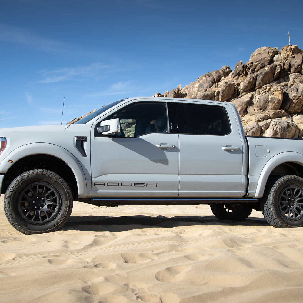 
                  
                    A white pickup truck with aggressive Roush styling and "Roush" lettering parked on sandy terrain, its 20-inch wheels glinting under the clear blue sky, rocky hills standing robust in the background.
                  
                