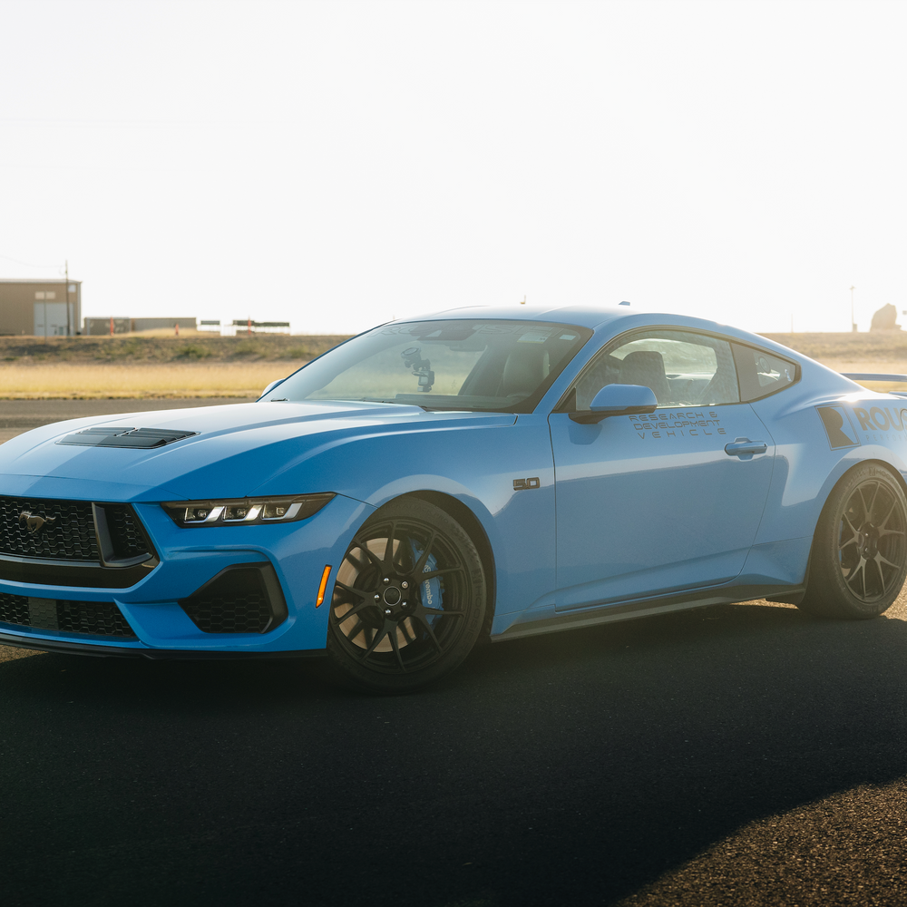 A blue sports car, featuring Roush Mustang Lowering Springs for an aggressive drop, gleams on a sunlit asphalt surface against an industrial backdrop.