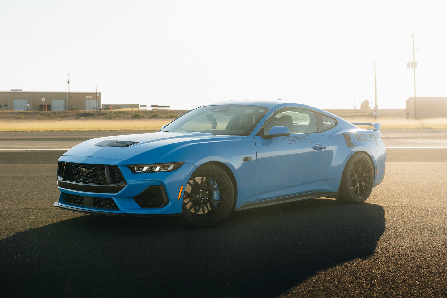 
                  
                    A blue sports car, featuring Roush Mustang Lowering Springs for an aggressive drop, gleams on a sunlit asphalt surface against an industrial backdrop.
                  
                