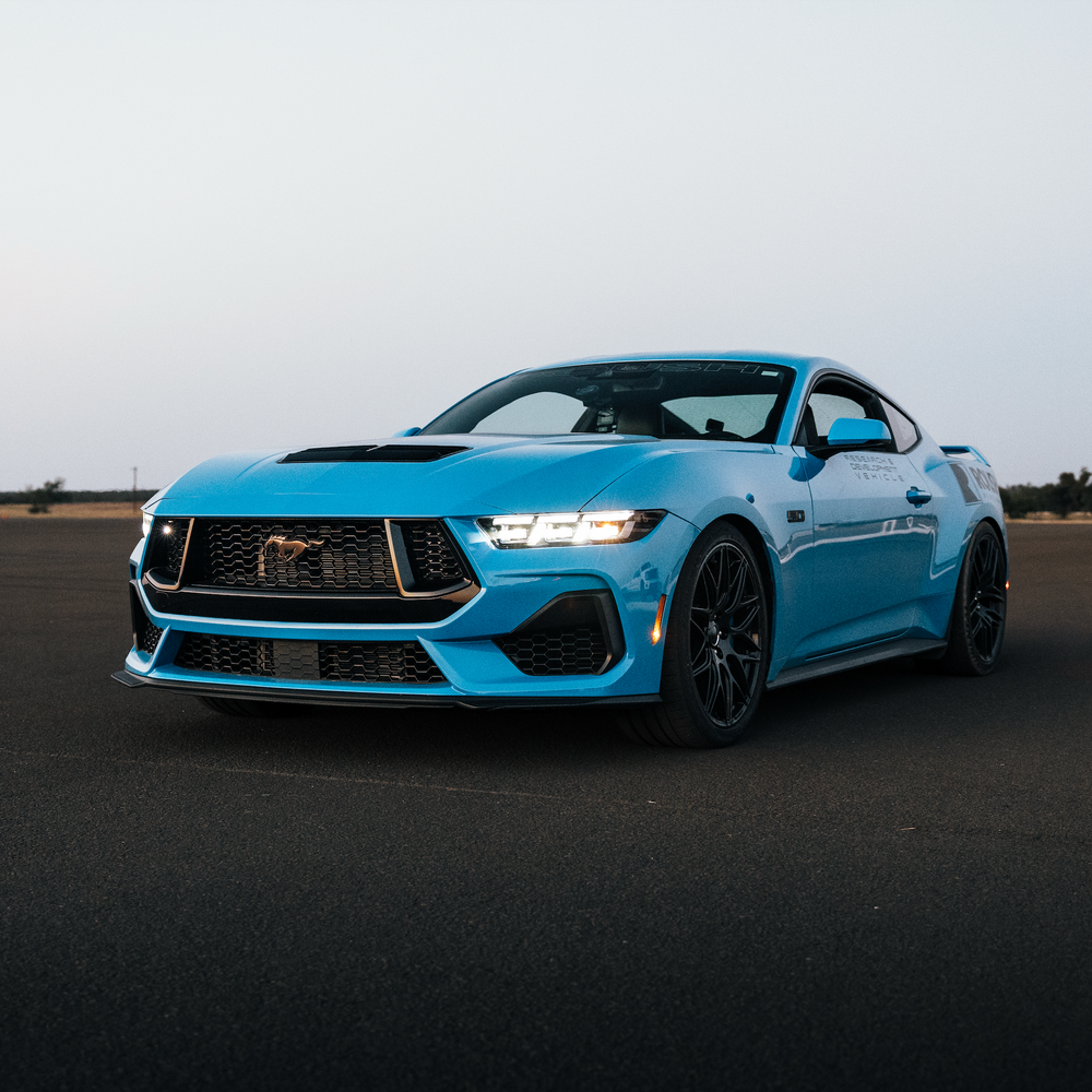 
                  
                    A blue sports car, equipped with aggressive drop and Roush Mustang Lowering Springs, is parked on a wide, empty asphalt surface under a clear sky.
                  
                