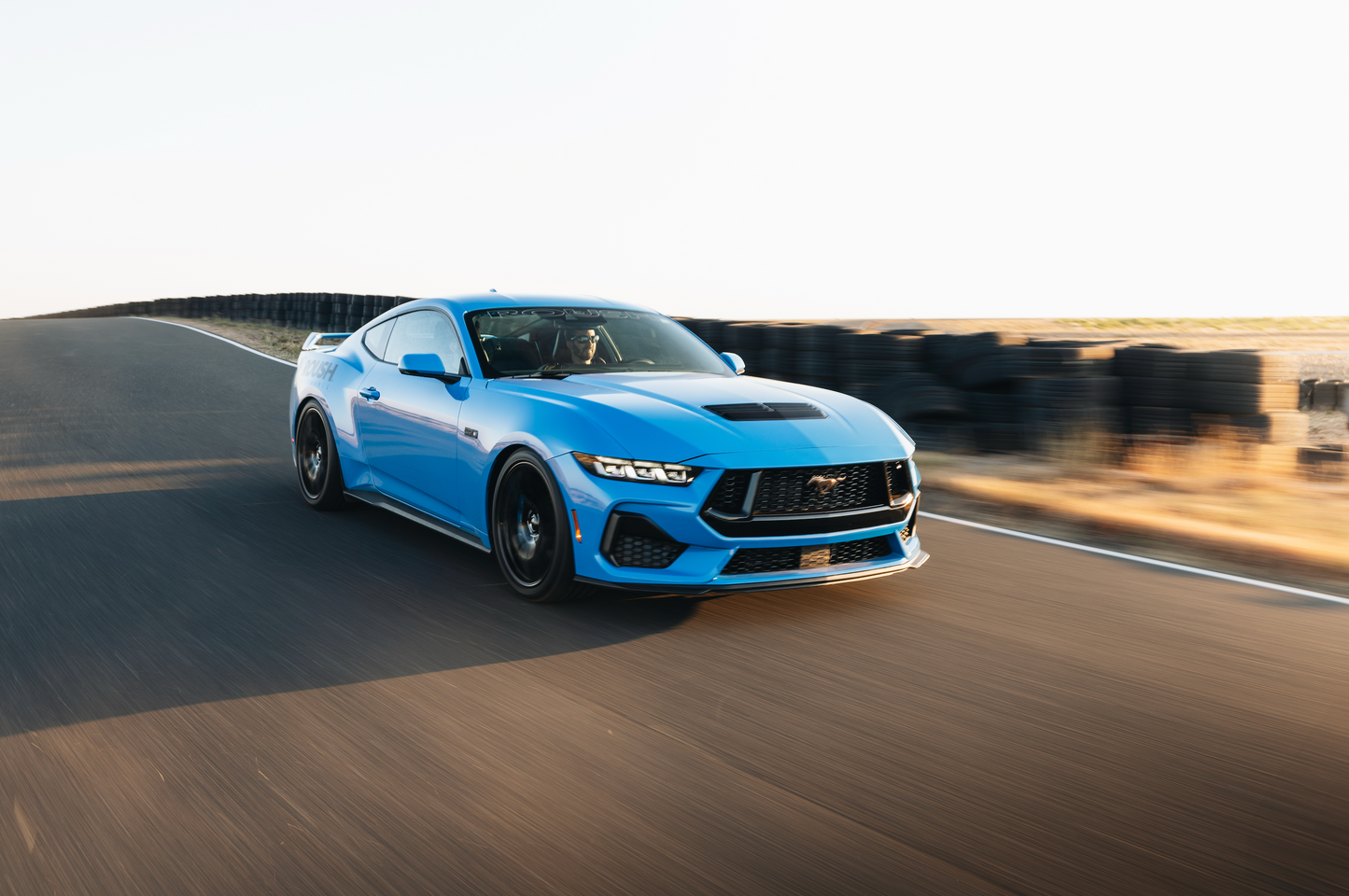 
                  
                    A blue sports car zips around the road track, its sleek profile accentuated by an aggressive drop from Roush Mustang Lowering Springs. Tire barriers stand in the background as it maneuvers with optimized spring rates for precise handling.
                  
                