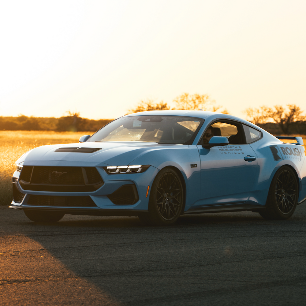 
                  
                    The Roush Mustang, featuring optimized spring rates, is a striking blue sports car parked on the road, its silhouette bold against the glow of a sunset-lit open field.
                  
                